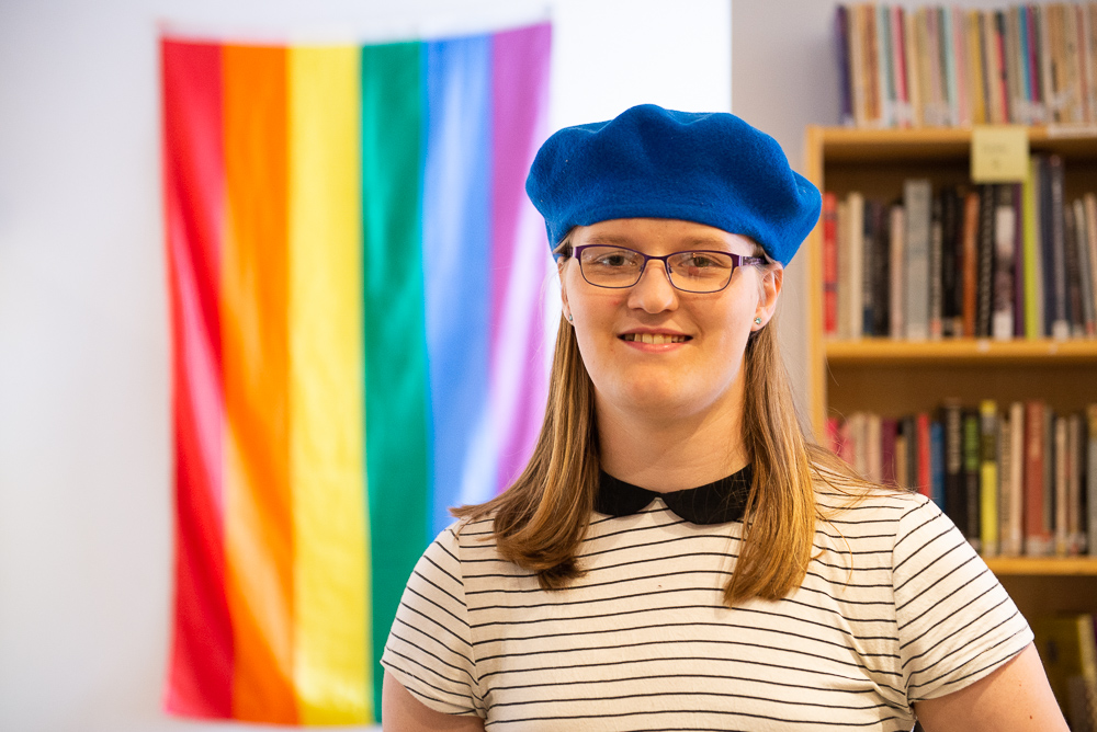 A photo of Penelope in a library standing in front of a rainbow flag wearing a blue beret.
