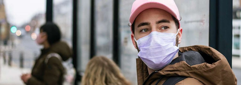 A teen wearing a pink mask and waiting to catch a bus.