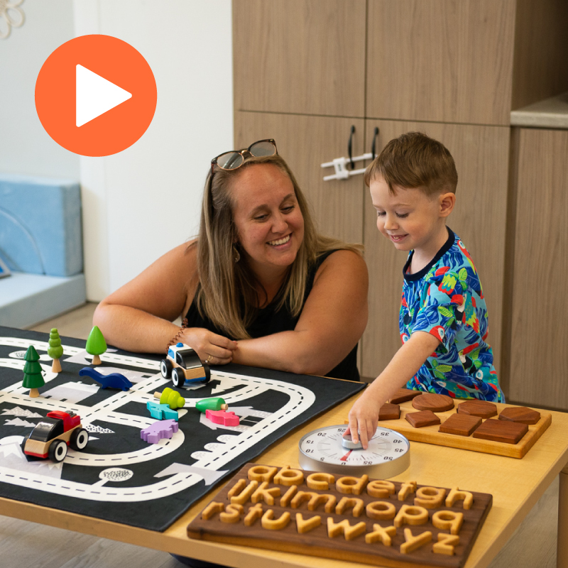 A smiling woman and a young boy are playing together at a table filled with educational toys. The table includes a toy road mat with small wooden cars and trees, a wooden alphabet puzzle, a shape-matching board, and a learning clock. The woman is watching attentively as the boy points at the clock. The scene takes place in a bright, child-friendly classroom or playroom. An orange play button icon is displayed in the top-left corner of the image.