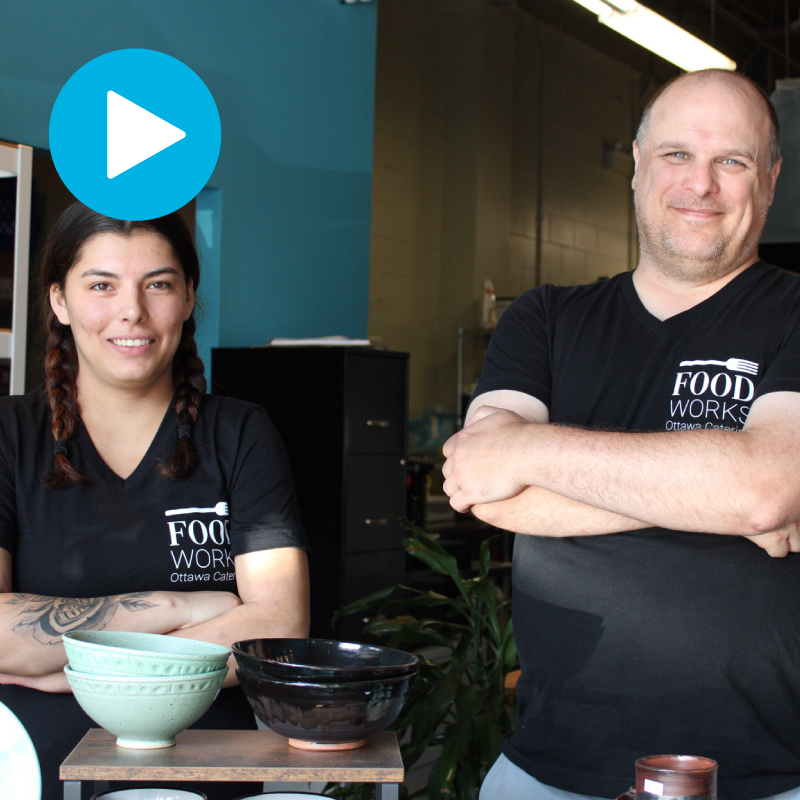 Two individuals, both wearing black FoodWorks Ottawa Catering t-shirts, stand proudly behind a display of handcrafted ceramic bowls. One person has braided hair and a visible tattoo, while the other has arms crossed and a welcoming smile. The setting appears to be a kitchen or workshop space with natural light and a relaxed atmosphere. A blue play button icon is displayed near the top-left corner of the image, suggesting video content. The image reflects a supportive and empowering environment focused on skill building and community.