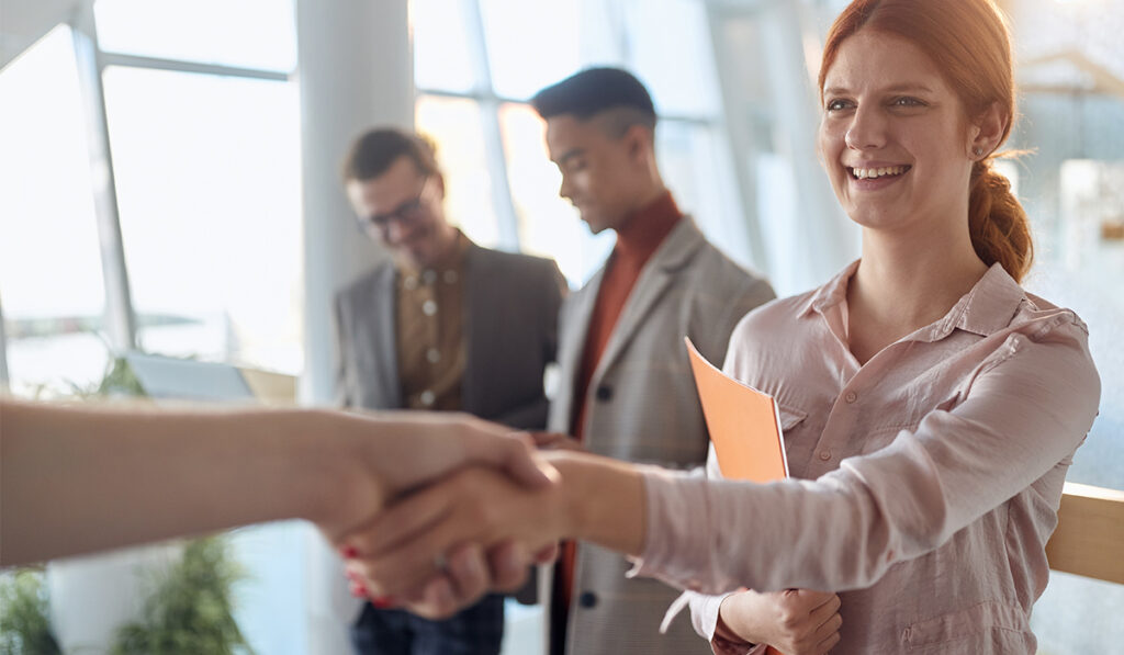 Smiling woman with red hair shakes hands while holding an orange folder, with two colleagues conversing in the background in a bright modern office.