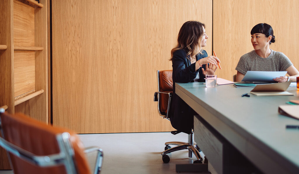 Two women sit at a conference table in a modern office, engaged in conversation and reviewing documents, with wooden walls and leather chairs in the background.
