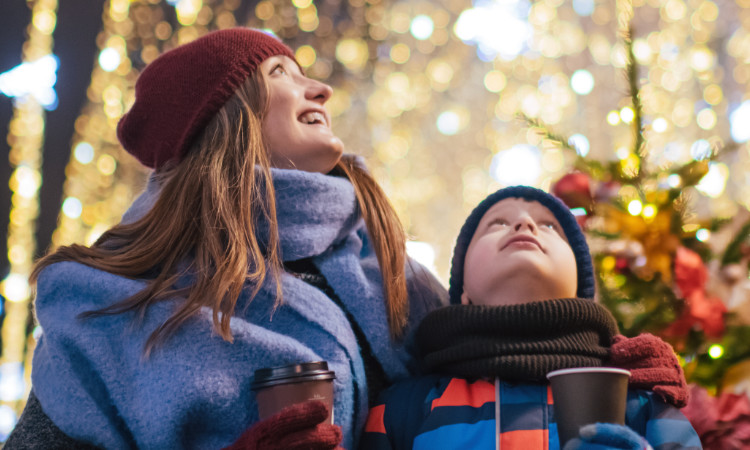 A photo of a Mom and her son outside looking up at holiday lights while enjoying some hot chocolate together.