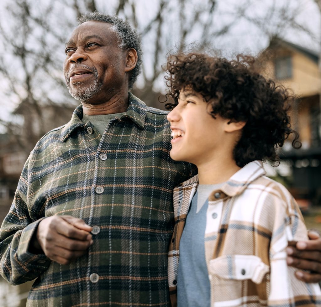 An older man and a young person smile together outdoors, standing with their arms around each other in a residential neighbourhood.